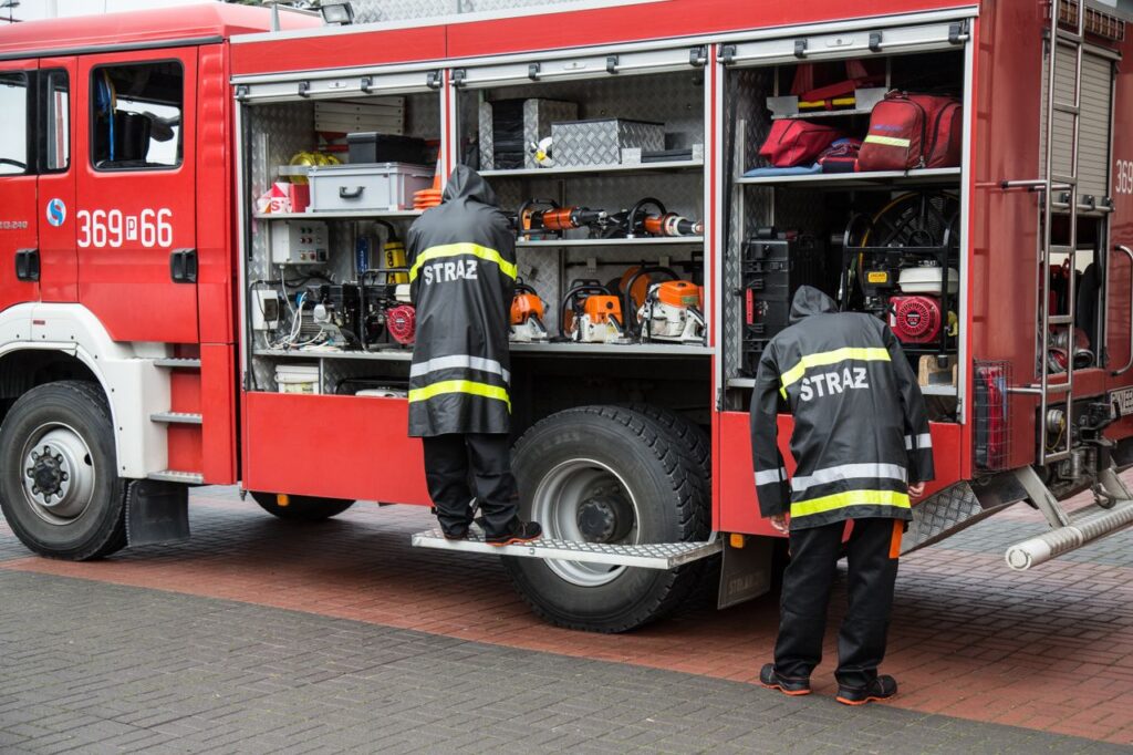 Firefighters in uniform organizing equipment on a firetruck with open compartments in an urban setting.